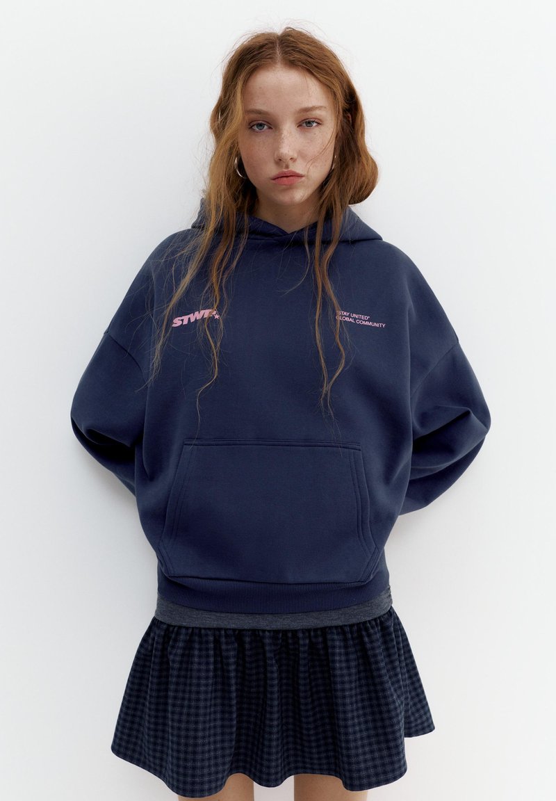 Giovane donna con lunghi capelli rossi che indossa una felpa con cappuccio blu navy con scritte rosa e una gonna scura a quadri, in piedi davanti a uno sfondo bianco.