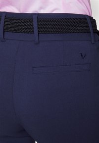 Zbliżenie tylnej kieszeni granatowych spodni z wyszywanym logo, czarnym plecionym paskiem i włożonym w pas różowym koszuli.