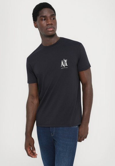 Tricou din bumbac bleumarin închis, cu mâneci scurte, guler rotund și un logo alb "AJX" pe pieptul stâng. Asortat cu blugi din denim albastră.