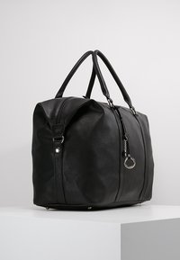 Sort faux læder duffel taske med lynlåslukning, to håndtag og en cirkulær metalaccent. Har en struktureret overflade og flad bund for stabilitet.