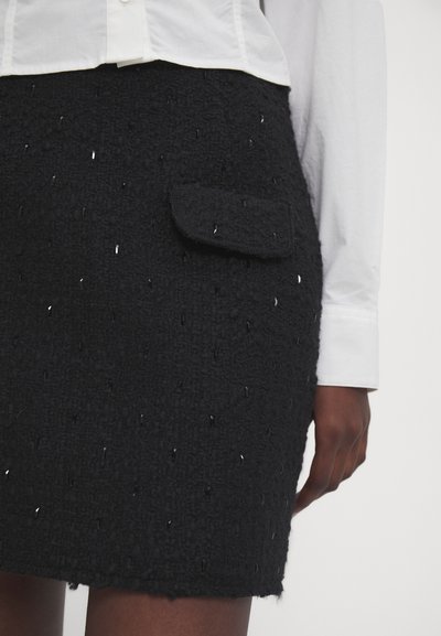 Jupe en tweed noir avec une surface texturée et des accents brillants, dotée d'une poche avant. Associée à une chemise blanche à boutons.