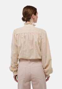 Blouse couleur crème avec col volé, manches bouffantes longues, tissu texturé et plis au dos, associée à un pantalon taille haute rose clair.
