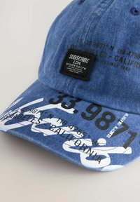 Blå denim baseballcap med en buet skygge, med sort og hvid tekst og grafik, samt et sort vævet mærke på forsiden.