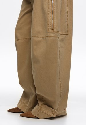 Pantalones kaki de pierna ancha con detalles de costuras, bolsillo lateral con cremallera, puños con botones que cubren zapatos marrones de punta afilada sobre fondo blanco.