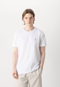 Tricou din bumbac alb cu mâneci scurte, având un mic broderie albastră "II" pe partea stângă a pieptului și un guler rotund.