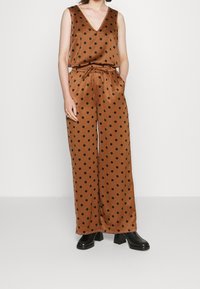 Combinaison en satin marron à pois noirs, avec un décolleté en V, une taille élastique et une coupe à jambes larges. Associée à des bottes à talons noires.