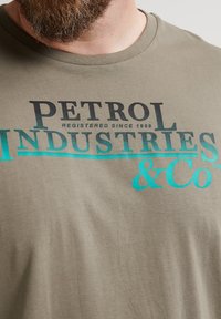 Olivově zelené bavlněné tričko s černým a tyrkysovým textem. Text obsahuje "PETROL" a "INDUSTRIES & Co" s poznámkou o registraci od roku 1989.