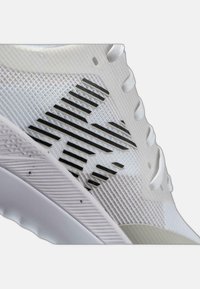 Zapatilla deportiva blanca con una parte superior texturizada que presenta rayas diagonales negras. Suela ligera y cordones, diseñada para el rendimiento y la comodidad.