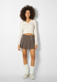 Krämskalad cardigan, brun rutiga veckad kjol med dubbelspänne, vita slouchiga strumpor och ljusa skor.