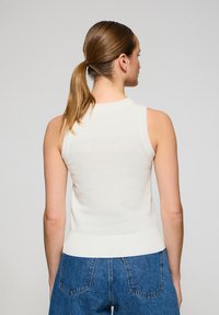 Top in maglia bianca senza maniche con orlo e aperture per le braccia a coste, indossato con jeans in denim blu a vita alta, che mostra un design pulito e semplice.
