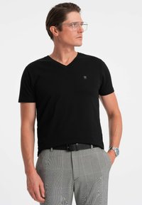 Sort v-hals t-shirt i et tekstureret stof, parret med mønstrede grå bukser. Bemærkelsesværdig logo detalje på venstre bryst og sølv ur på håndleddet.