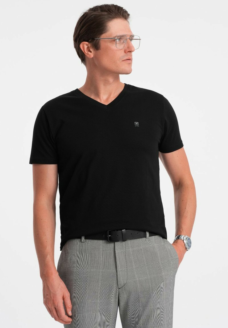 T-shirt nera a scollo a V in un tessuto texture, abbinata a pantaloni grigi con motivo. Dettaglio del logo ben visibile sul petto sinistro e orologio argentato al polso.