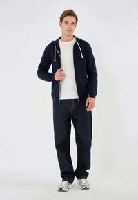 Marineblå zip-up hættetrøje med hvide snore, parret med mørkeblå jeans og grå sportsko. Modellen står foran en ensfarvet baggrund.