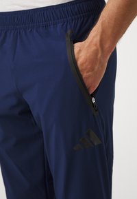 Marineblauwe sportbroek gemaakt van gladde stof, met een zwarte ritszak en een zwart logo op het onderbeen. Inclusief elastische tailleband.
