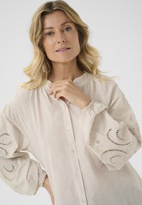 Chemise beige clair à boutons en coton avec des détails en dentelle élaborés sur les manches et des poignets froncés. Texture lisse ; coupe décontractée.