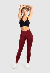Zwarte sportbh met ruchingdetail, bordeauxrode high-waisted leggins en witte sportschoenen op een grijze achtergrond.