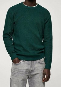 Homme portant un pull vert foncé à manches longues et un jean gris clair, une main dans la poche, debout devant un fond uni.