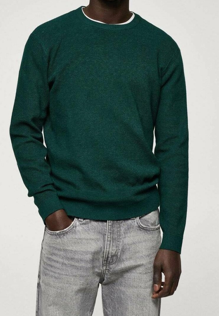 Homme portant un pull vert foncé à manches longues et un jean gris clair, une main dans la poche, debout devant un fond uni.