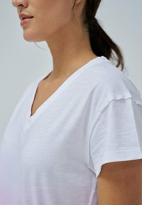 Camiseta blanca de manga corta y escote en V, confeccionada en una tela ligera y texturizada, con detalles sin terminar en las mangas y el escote.