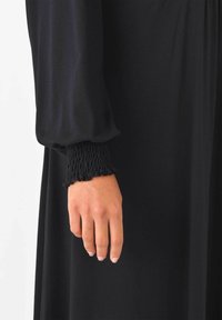 Robe noire avec de longues manches bouffantes. Les poignets présentent des détails élastiques texturés froncés. Le tissu lisse tombe bien depuis la taille.