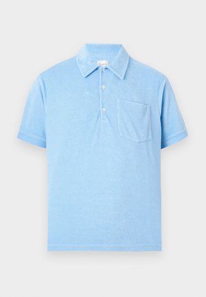 Mand stående med hænderne i lommerne iført lyseblå kortærmet polo, mørkeblå denimshorts og brune lædersandaler.