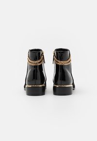 Botas de tobillo de charol negro con detalles en cadena dorada, cremalleras laterales y puntera redondeada. Tacón negro resistente y costuras decorativas.