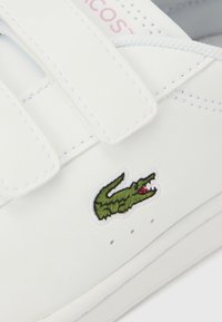 Λευκό συνθετικό αθλητικό παπούτσι με λουράκια βέλκρο, διαθ έτει κέντημα λογότυπου με πράσινο κροκόδειλο. Λευκή επιφάνεια με διακριτικές λεπτομέρειες ραφής.