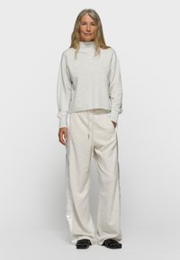 Frau mit langen grauen Haaren, die einen hellbeigen Rollkragenpullover und passende weit geschnittene Hosen mit Satin-Seitenstreifen trägt, schwarze Schuhe.