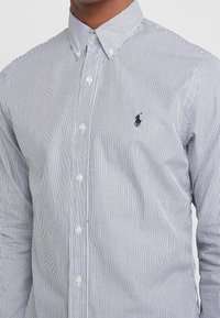 Mężczyzna w białej koszuli z długim rękawem w cienkie pionowe czarne paski oraz z czarnym haftowanym logo polo na piersi.