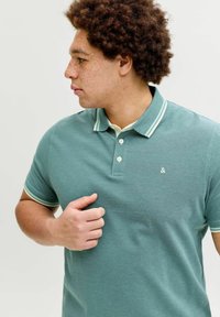 Νέος άνδρας με σγουρά μαλλιά και φακίδες, φορώντας μια πετρόλ πόλο μπλούζα με λευκή επένδυση, κοιτάζει προς τα αριστερά του, με το χέρι κοντά στο στήθος.