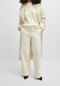 Femme portant un pantalon ample crème, un pull en maille crème et un manteau texturé beige, se tenant avec les mains dans les poches et des chaussures à talons noirs.