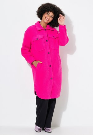 Abrigo de forro polar en rosa brillante con collar, dos bolsillos frontales y botones negros. Llevado sobre pantalones negros y zapatos plateados brillantes. Diseño con paneles.