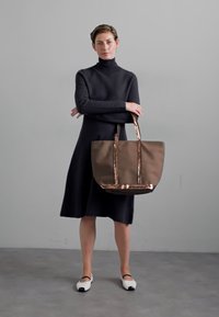 Sac fourre-tout marron avec des poignées en métal rose doré, texture lisse, grande forme rectangulaire. Porté avec une robe noire ajustée et des chaussures contrastantes.