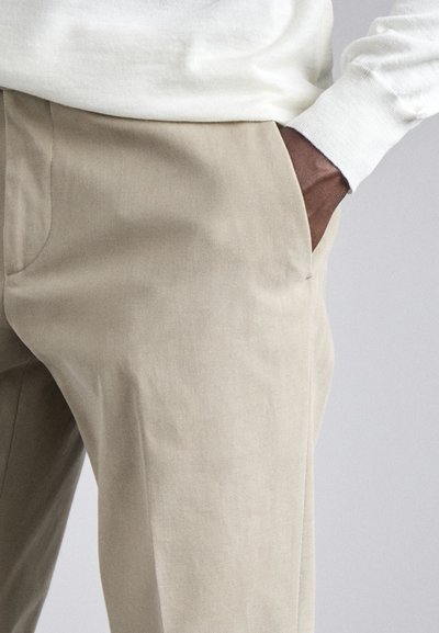 Beige Baumwollhosen mit glatter Textur, seitlichen Taschen und entspannter Passform, kombiniert mit einem hellen Strickpullover.