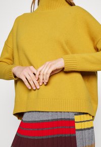 Kobieta w musztardowym swetrze i plisowanej spódnicy w szare, czerwone, bordowe i żółte paski, delikatnie złożone ręce na talii.