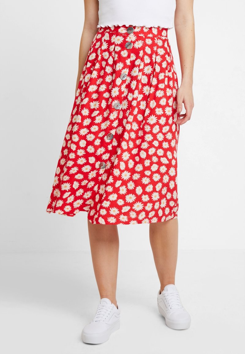 Jupe rouge à imprimé marguerites blanches avec boutons sur le devant, portée avec des baskets blanches et un haut blanc court, vue de la taille aux pieds.