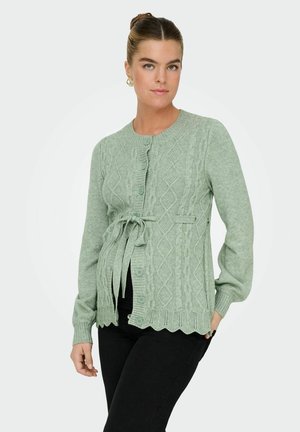 Cardigan a maglia verde chiaro con motivo a trecce e orlo scallop, dotato di vita legata e chiusure a bottone, indossato sopra pantaloni neri.