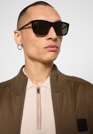 Giovane con capelli corti che indossa occhiali da sole rettangolari neri, orecchino a cerchio d'argento, maglia beige con zip e giacca marrone con collo a coste.