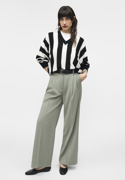 Γυναίκα που στέκεται φορώντας πουλόβερ με μαύρες και λευκές κάθετες ρίγες, ανοιχτό γκρι παντελόνι με φαρδιά μπατζάκια, μαύρη ζώνη και μαύρα φλατ παπούτσια.