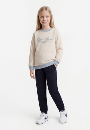 Ξανθιά κοπέλα που χαμογελά, φοράει μπεζ φούτερ με μπλε περιθώριο, σκούρες μπλε παντελόνες και λευκά παπούτσια Nike, στέκεται με το ένα χέρι στην τσέπη.