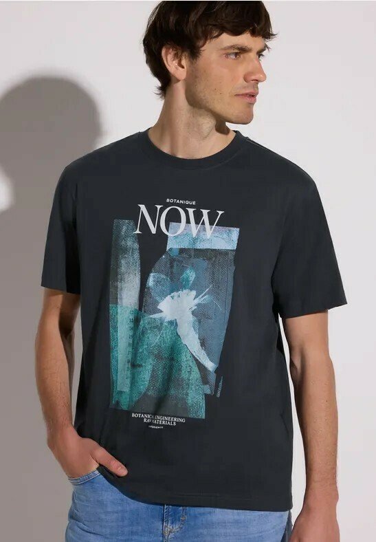 Homme portant un T-shirt graphique noir avec un design abstrait bleu-vert et le texte « BOTANIQUE NOW » et un jean bleu clair, regardant vers la droite.