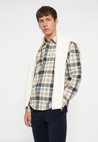 Geruit shirt in crème, marineblauw en bruin met knoopsluiting aan de voorkant; gecombineerd met een textuurwit sweater over de schouders; marineblauwe broek.