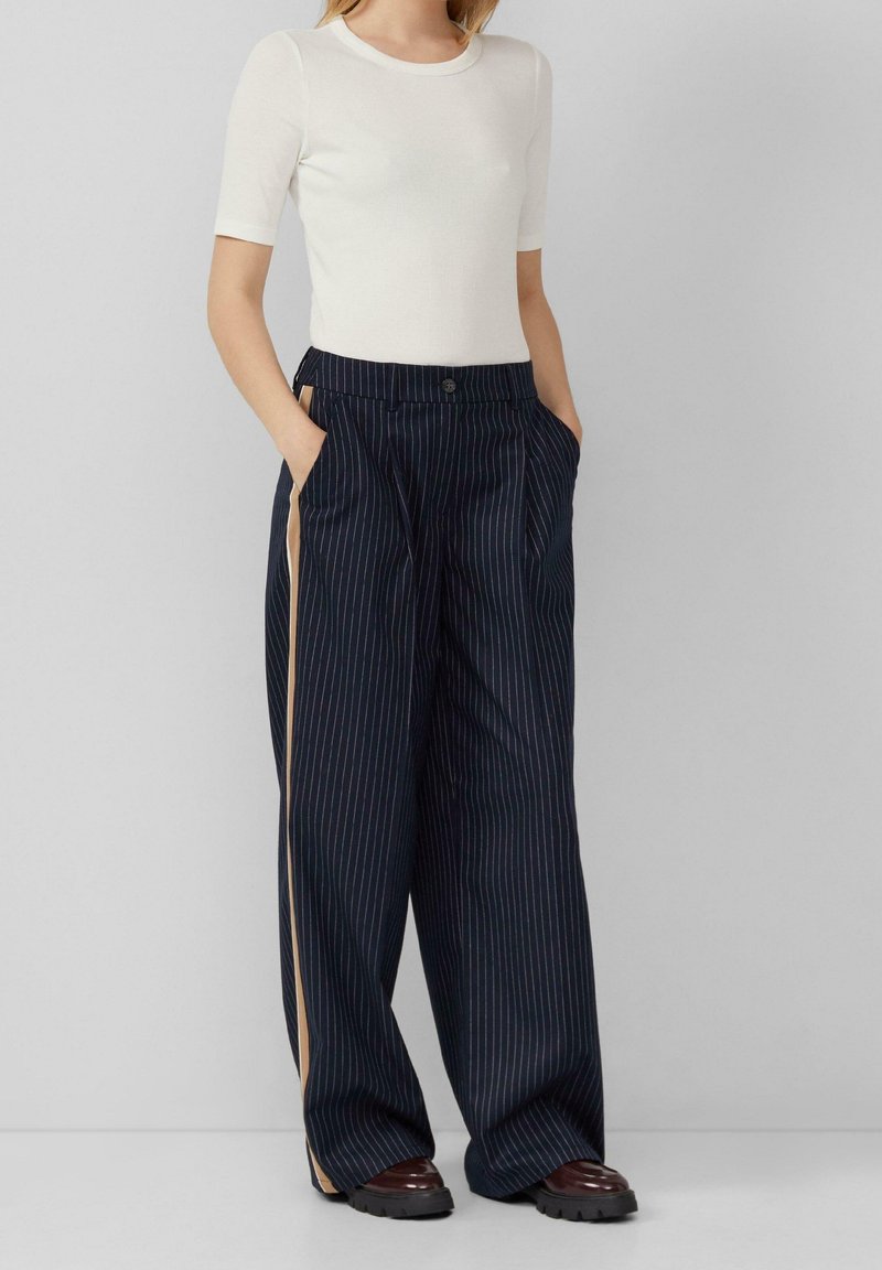 Femme portant un haut blanc à manches courtes, pantalon large bleu marine à fines rayures avec une bande latérale beige, mains dans les poches et chaussures épaisses marron foncé.
