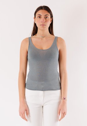 Donna con lunghi capelli scuri che indossa una canotta grigia e pantaloni bianchi, in piedi contro uno sfondo bianco uniforme.