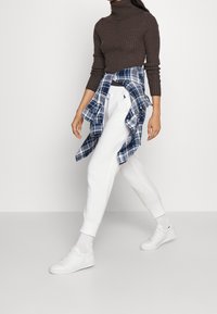 Brun randig polotröja, vita sweatpants med uppvikta anklar och vita sneakers. Blå rutiga skjorta knuten runt midjan.