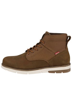 Botas de tobillo marrones con cordones, parte superior de ante, paneles laterales de lona, suela de goma blanca y detalles de costura. Presenta una etiqueta con el logo en el lateral.