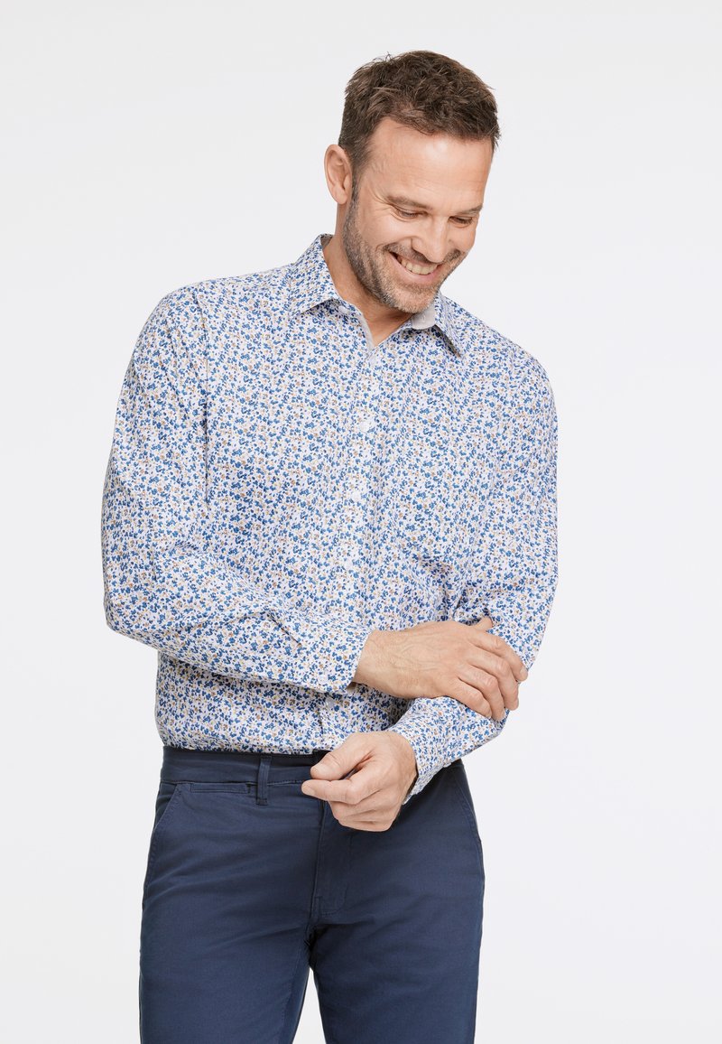Camicia a maniche lunghe con base bianca caratterizzata da un motivo floreale blu, tessuto intrecciato e dettagli con bottoni. Abbinata a pantaloni blu navy.
