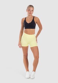 Sujetador deportivo negro con diseño de escote en v y pantalones cortos amarillos de cintura alta. Modelo con zapatillas blancas. Fondo limpio, enfocado en el conjunto.