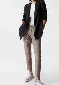 Femme portant un blazer noir, un top rayé blanc, un pantalon cropped à carreaux, et des baskets blanches, debout avec les mains dans les poches contre un fond blanc.