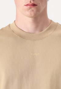 Beige Rundhals-T-Shirt aus glattem Stoff, mit einem dezenten, in der gleichen Farbe auf der Brust geprägten "HUGO"-Logo.